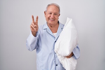 Senior man with grey hair wearing pijama hugging pillow smiling looking to the camera showing fingers doing victory sign. number two.