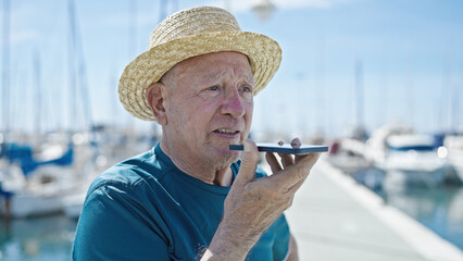 Senior grey-haired man tourist wearing summer hat sending voice message by smartphone at port