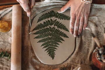 Female sculptor making clay pottery in a home workshop.Making an imprint of a fern leaf on a clay...