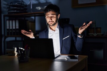 Handsome latin man working at the office at night clueless and confused expression with arms and hands raised. doubt concept.