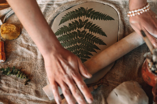 Female sculptor making clay pottery in a home workshop.Making an imprint of a fern leaf on a clay plate,hands close-up.Small business,entrepreneurship,hobby, leisure,sustainability concept.