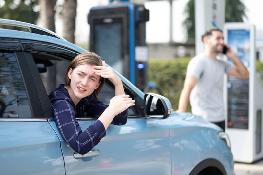 White woman sitting in her car driving side, park at electrical fluel engine spot looking back with concfuse or looking for someone, her boyfriend add fluel and talking on smartphone.