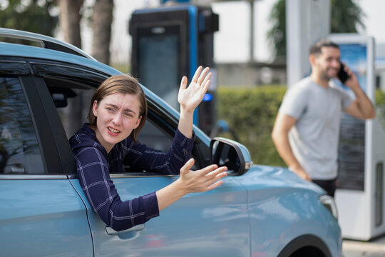 White woman sitting in her car driving side, park at electrical fluel engine spot looking back with aggresssive emotion or confuse with other, her boyfriend add fluel and talking on smartphone.