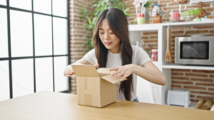 Young chinese woman unpacking cardboard box sitting on table with serious expression at dinning room