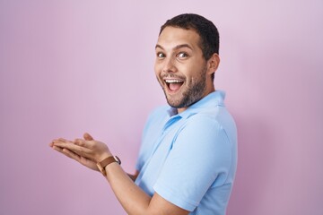 Hispanic man standing over pink background pointing aside with hands open palms showing copy space, presenting advertisement smiling excited happy