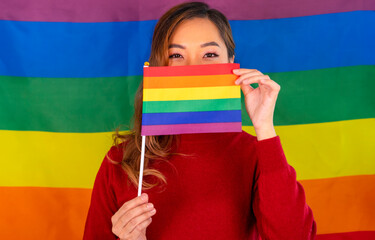 asian woman hands holding lgbt rainbow flag cover her mouth the eyes looking at camera, background rainbow stripes, concept of lgbtq community, protest,movement,claim,rights,freedom