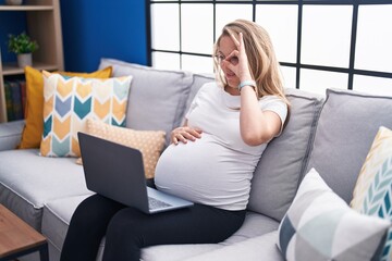 Young pregnant woman sitting on the sofa at home using laptop smiling happy doing ok sign with hand on eye looking through fingers