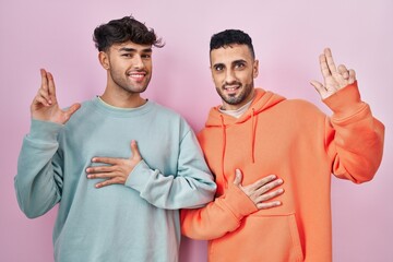 Young hispanic gay couple standing over pink background smiling swearing with hand on chest and...