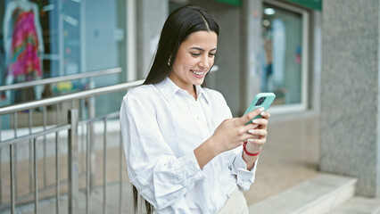 Young beautiful hispanic woman using smartphone smiling at street