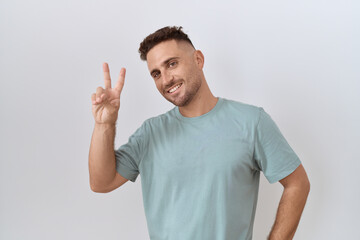 Hispanic man with beard standing over white background smiling looking to the camera showing fingers doing victory sign. number two.