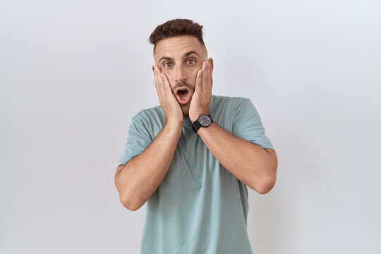 Hispanic man with beard standing over white background afraid and shocked, surprise and amazed expression with hands on face