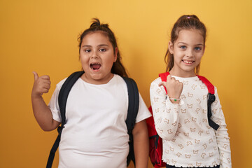 Two little girls friends wearing student backpack pointing thumb up to the side smiling happy with open mouth