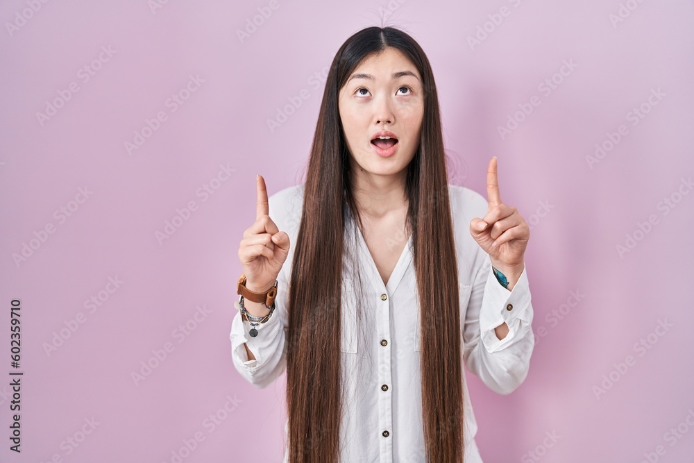 Sticker Chinese young woman standing over pink background amazed and surprised looking up and pointing with fingers and raised arms.