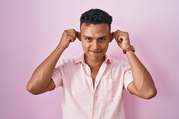 Young hispanic man standing over pink background smiling pulling ears with fingers, funny gesture....