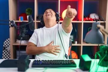 Young hispanic man playing video games laughing at you, pointing finger to the camera with hand over body, shame expression