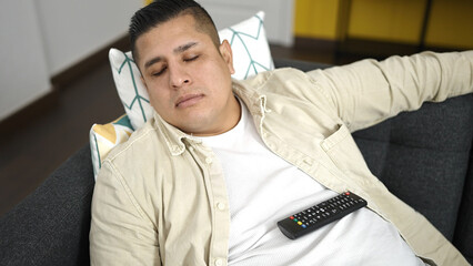 Young hispanic man holding tv remote control lying on sofa sleeping at home