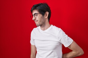 Young hispanic man standing over red background suffering of backache, touching back with hand, muscular pain