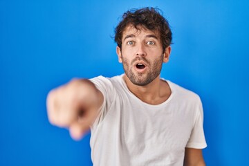 Hispanic young man standing over blue background pointing with finger surprised ahead, open mouth amazed expression, something on the front