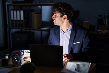 Hispanic young man working at the office at night looking to side, relax profile pose with natural face with confident smile.