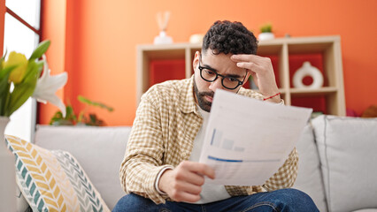 Young hispanic man reading document sitting on sofa at h