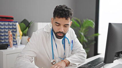 Young hispanic man doctor using computer writing notes at clinic