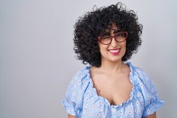 Young brunette woman with curly hair wearing glasses over isolated background winking looking at...