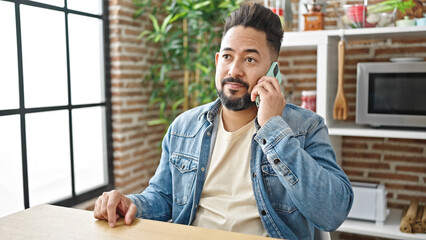 Young latin man talking on smartphone sitting on table at dinning room