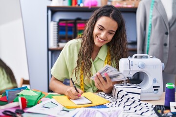 Young beautiful hispanic woman tailor holding dollars writing on notebook at clothing factory