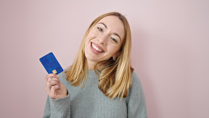 Young blonde woman smiling confident holding credit card over isolated pink background