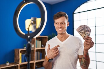 Caucasian man recording vlog tutorial with smartphone at home holding money smiling happy pointing with hand and finger
