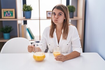 Obraz na płótnie Canvas Young hispanic woman drinking glass of orange juice puffing cheeks with funny face. mouth inflated with air, catching air.