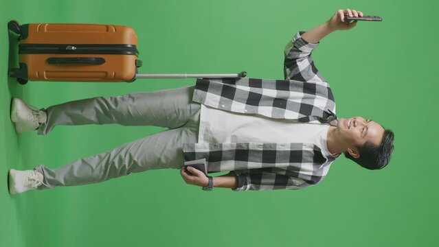 Full Body Of Asian Male Traveler With Luggage And Passport Smiling And Taking Photo On Smartphone While Standing In The Green Screen Background Studio
