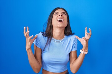 Fototapeta premium Brunette young woman standing over blue background crazy and mad shouting and yelling with aggressive expression and arms raised. frustration concept.