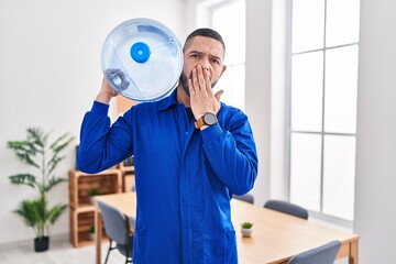 Hispanic service man holding a gallon bottle of water for delivery covering mouth with hand,...