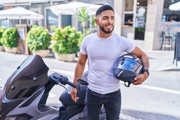 Young latin man holding helmet standing by motorbike at street