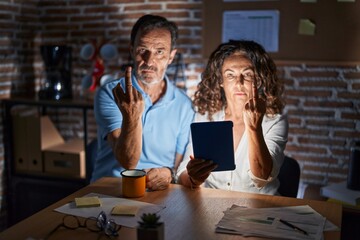Middle age hispanic couple using touchpad sitting on the table at night showing middle finger,...