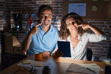 Middle age hispanic couple using touchpad sitting on the table at night smiling cheerful showing and pointing with fingers teeth and mouth. dental health concept.