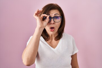 Middle age hispanic woman standing over pink background doing ok gesture shocked with surprised face, eye looking through fingers. unbelieving expression.