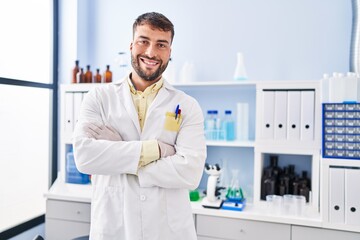 Young hispanic man wearing scientist uniform standing with arms crossed gesture at laboratory