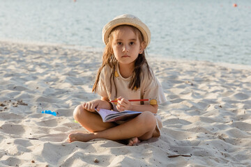 A little cute girl sits on the beach on vacation and draws on paper with colored pencils. Family on a summer weekends. Little girl drawing. Happy summer vacation concept