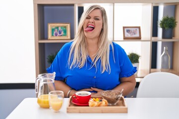 Obraz na płótnie Canvas Caucasian plus size woman eating breakfast at home sticking tongue out happy with funny expression. emotion concept.