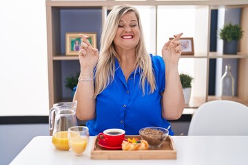 Obraz na płótnie Canvas Caucasian plus size woman eating breakfast at home gesturing finger crossed smiling with hope and eyes closed. luck and superstitious concept.