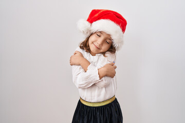 Little hispanic girl wearing christmas hat hugging oneself happy and positive, smiling confident. self love and self care