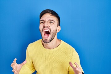 Hispanic man standing over blue background crazy and mad shouting and yelling with aggressive expression and arms raised. frustration concept.