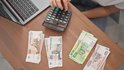 Young blonde woman counting ruble banknotes using calculator at home