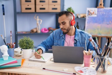Young hispanic man artist using laptop and headphones drawing on notebook at art studio