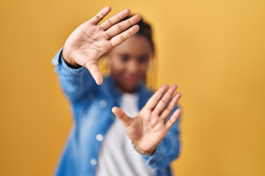 African american woman with braids standing over yellow background doing frame using hands palms and fingers, camera perspective