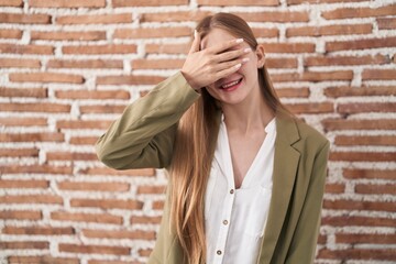 Young caucasian woman standing over bricks wall background smiling and laughing with hand on face covering eyes for surprise. blind concept.