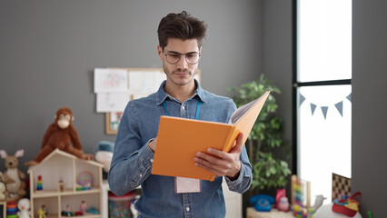 Young hispanic man preschool teacher reading book with relaxed expression at kindergarten