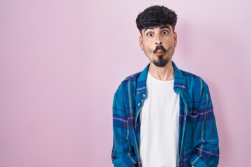 Young hispanic man with beard standing over pink background making fish face with lips, crazy and comical gesture. funny expression.